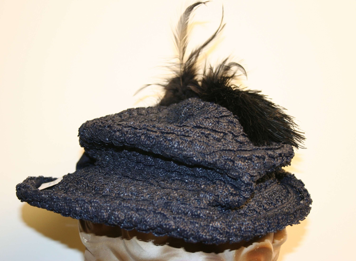 En mørk blå stråhatt med fjær til kvinne. Sekundære svarte fjær, ripsbånd rundt pullen i hvitt og svart.