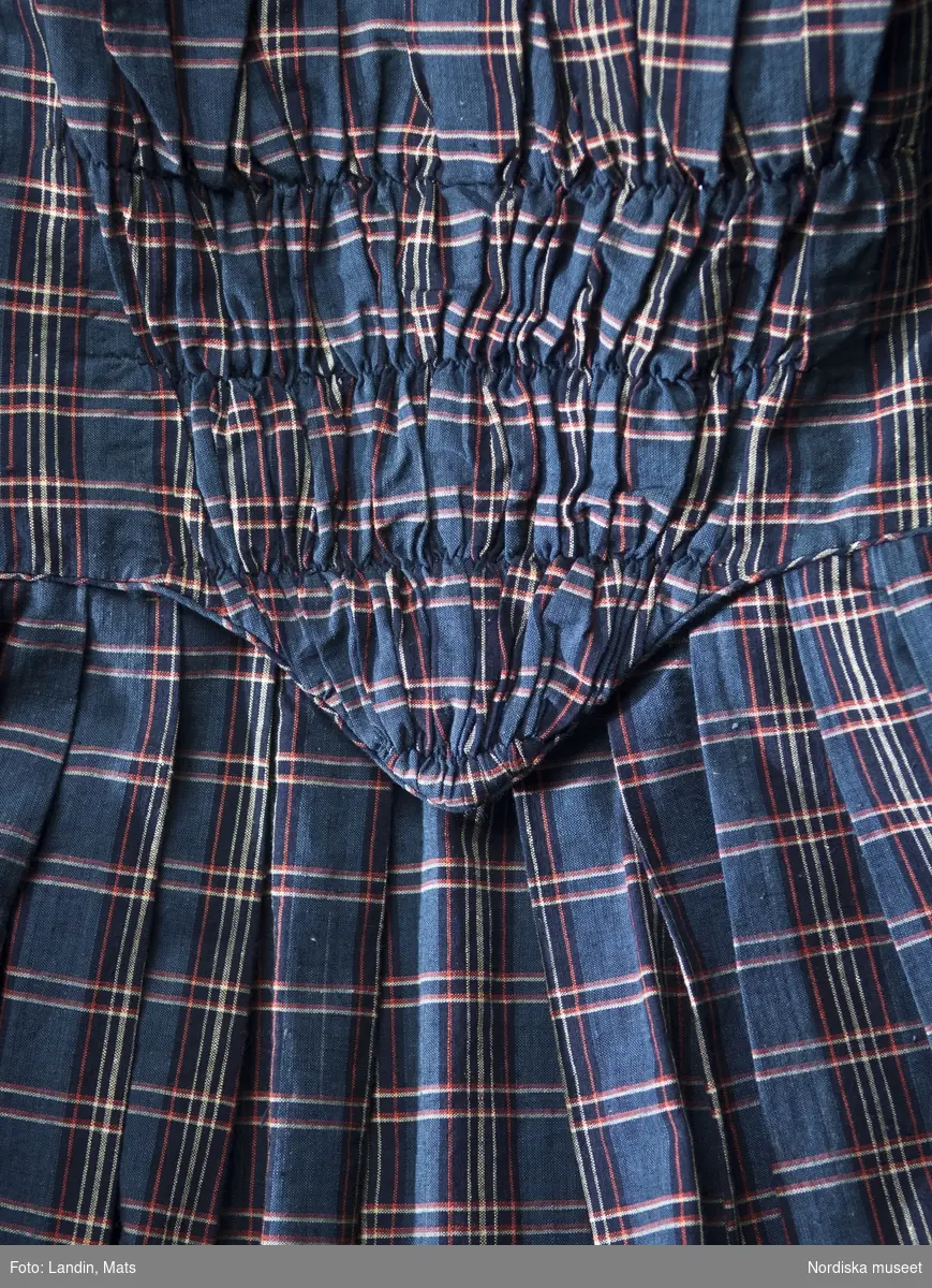 Högtids klänning, 1860-talet
Värmland, Kristinehamn 
Sömmerska, Bomull

Klänning av hemvävt rutigt bomullstyg. V-formad besättning med rynkor över framstycket, bakstycket är rynkat och draperat i ryggen. Baktill en skörtflik. Lång ärm med knäppt sprund, garnering i form av en kort överärm som är kantad med snedslå av tyget och liten kokard. I ärmsöm, midjesöm och kring framkanter passepoaler (insydd dekorkant ofta i avvikande färg i en söm) av tyget. Har tillhört Clara Westman (1845-1930), f. Berggren, dotter till brukspatron Magnus Theodor Berggren  vid Niklasdams bruk , Kristinehamn, Värmland. Gift 1869 med Robert Westman, ekonomichef i Järnvägsstyrelsen. Clara använde klänningen innan hon gifte sig.