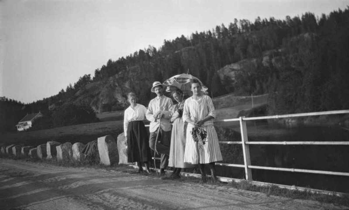 Tre damer og en mann står på en bro. Den ene damen med parasoll.