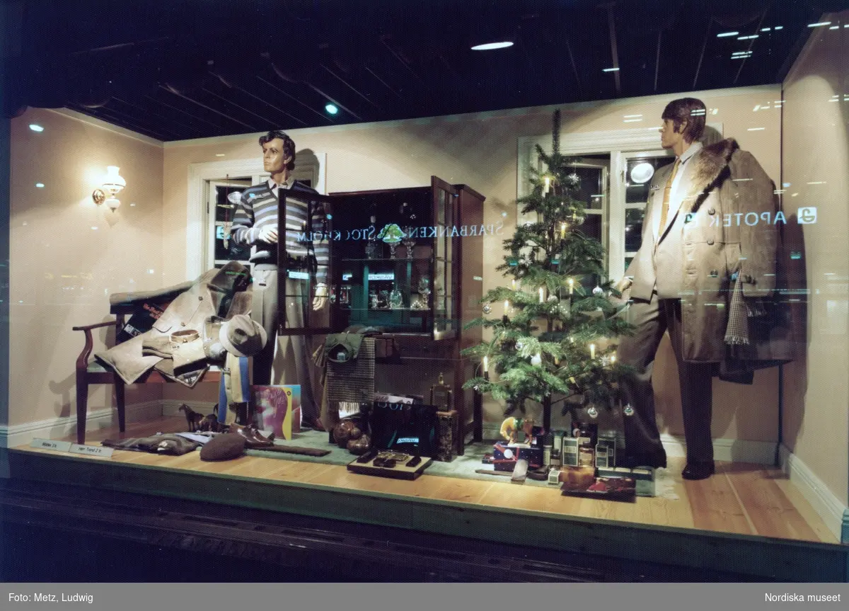 Skyltfönster på Nordiska Kompaniet. Julskyltning. Herrkläder - kostym, rockar, tröjor, skjortor, accessoarer och parfymer i vardagsrumsdekor med juldekorationer.