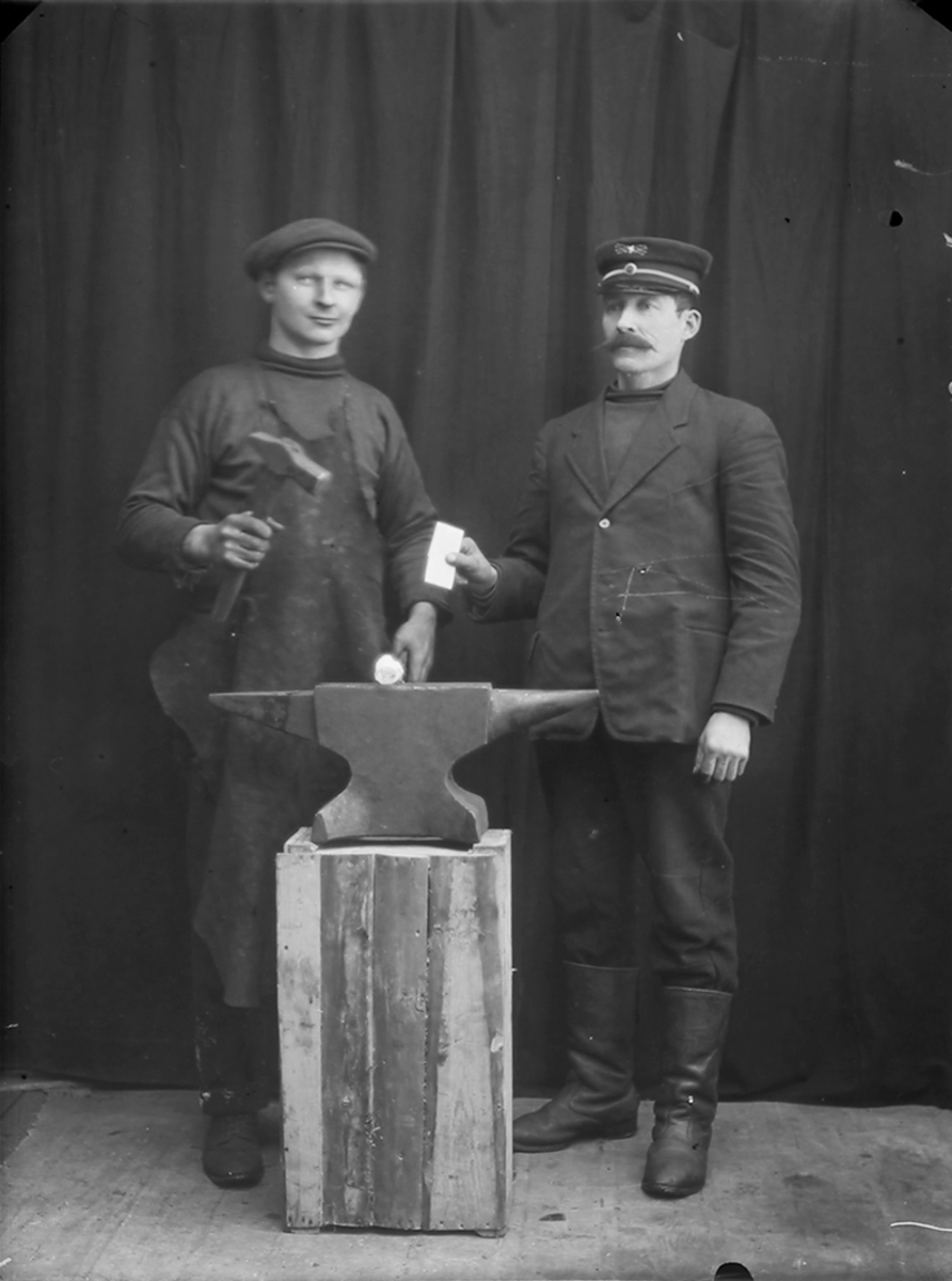 Portrett. 2 menn ved en ambolt. Den ene har en hammer. Den andre har uniformslue på hodet. Muligens lensmann eller jernbanefunksjonær.Bildet er tatt på Reine i Lofoten.
