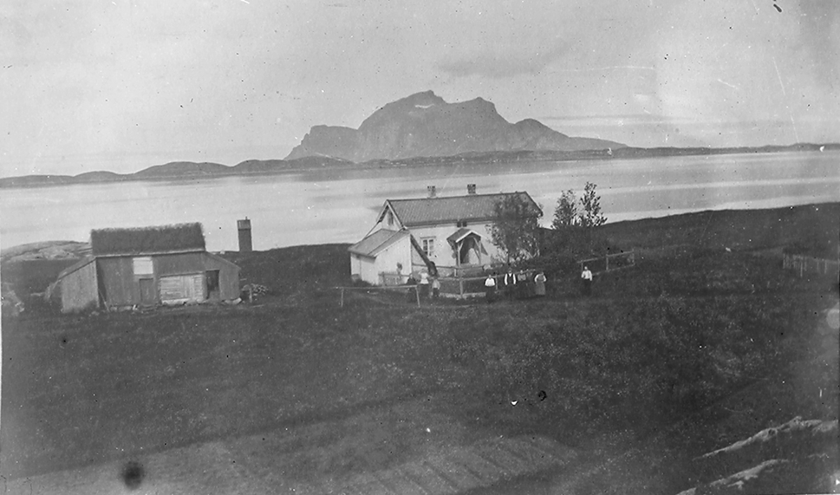 Landskapsbilde, gård foran, våningshus og fjøs. 9 mennesker står foran våningshuset. I bakgrunnen sees sjøen og fjell, mulig øy?