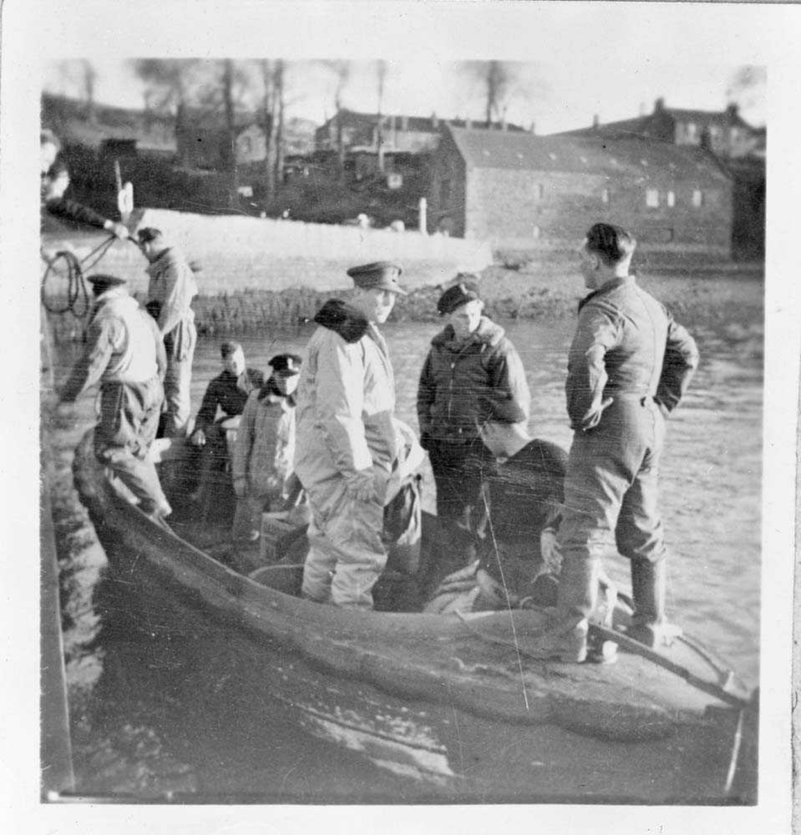 Nordahl Grieg står midt i bildet  i lys flydress med mørk krage og hærens uniformslue. Til høyre for ham står muligens Jon O. Roe i mørk flygerjakke og marinens uniformslue. Båten de står i skal frakte dem ut til et Catalina-fly.
