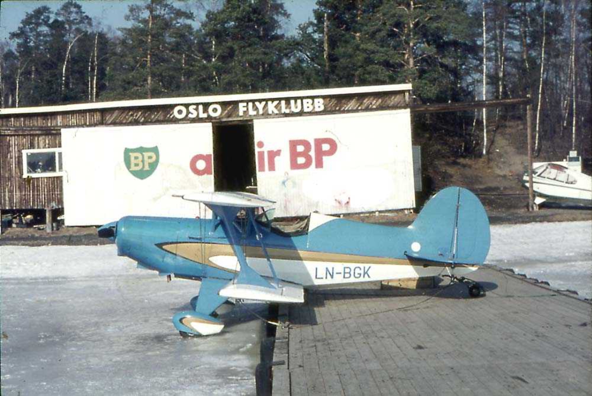 Ett fly som står med forparten på et isete vann og bakparten på ei trekai., EAA Acro Sport, LN-BGK. Bygning og et fly i bakgrunnen.