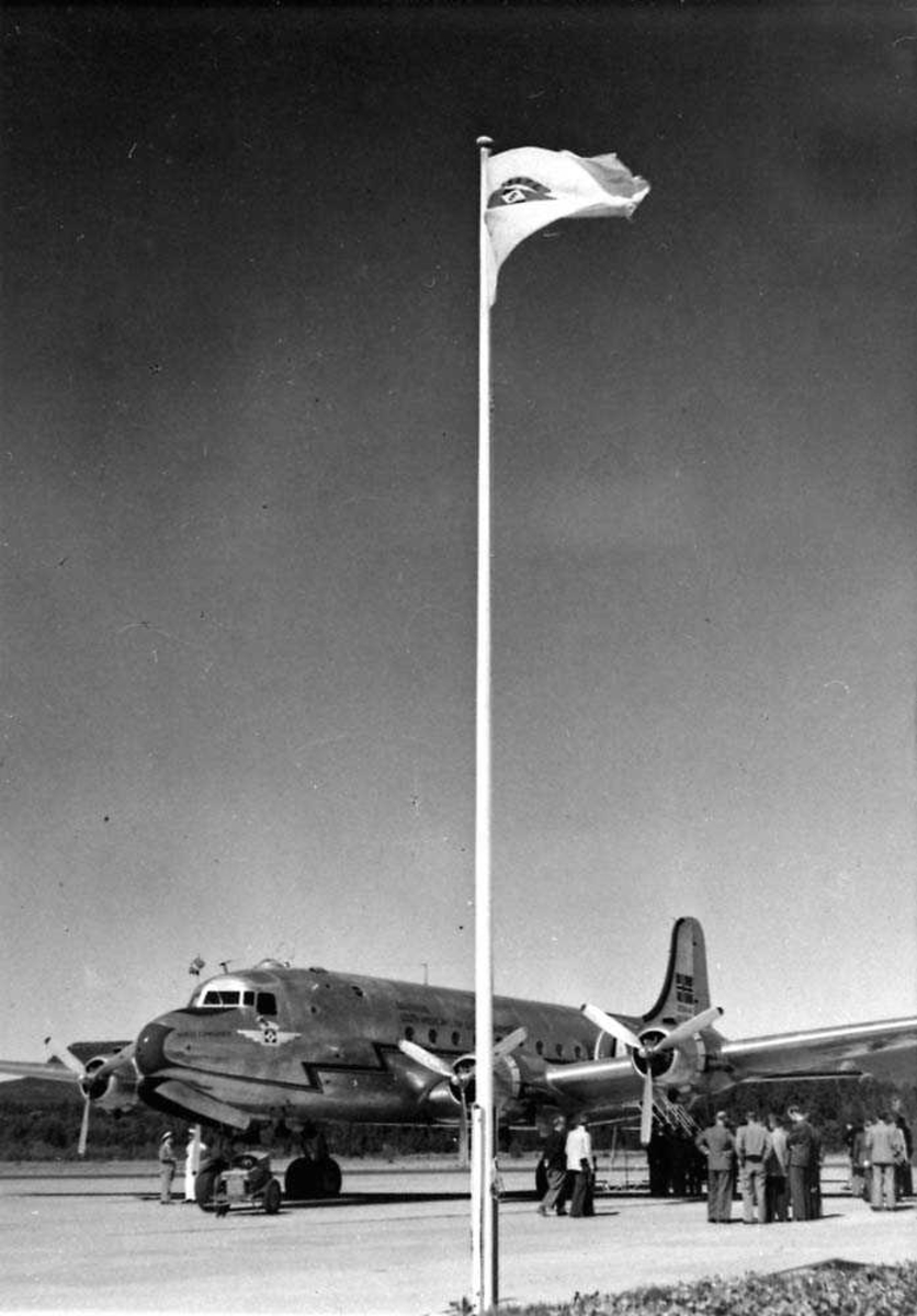 Ett fly på bakken. Douglas DC-4/C-54 "Norse Comander" LN-PAW fra Braathens SAFE. Flere personer rundt flyet. Flaggstang med Braathens SAFE flagg i forgrunnen.