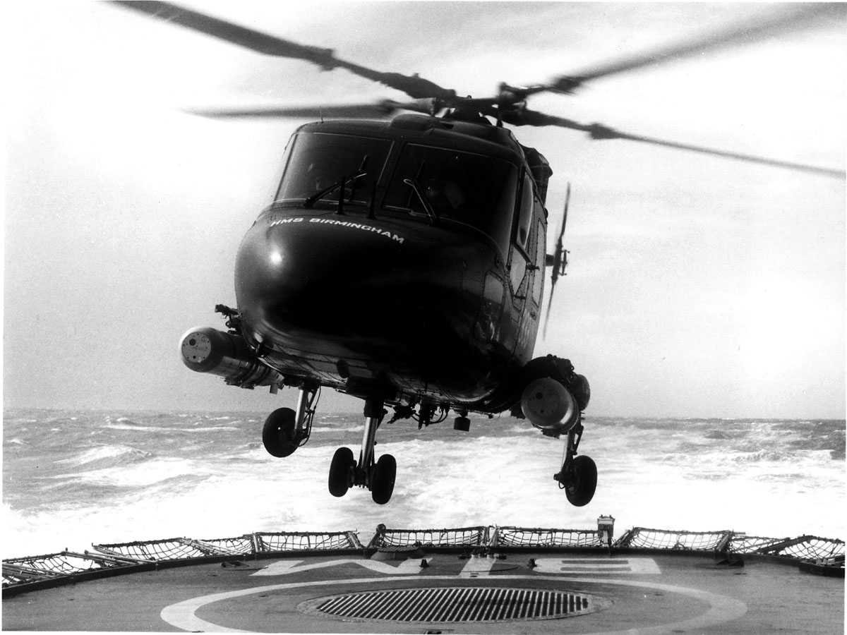Ett helikopter i luften, like over helipad/skipsdekk. 2 torpedoer hengende på hver sin side. Westland Lynx (ukjent Mk)