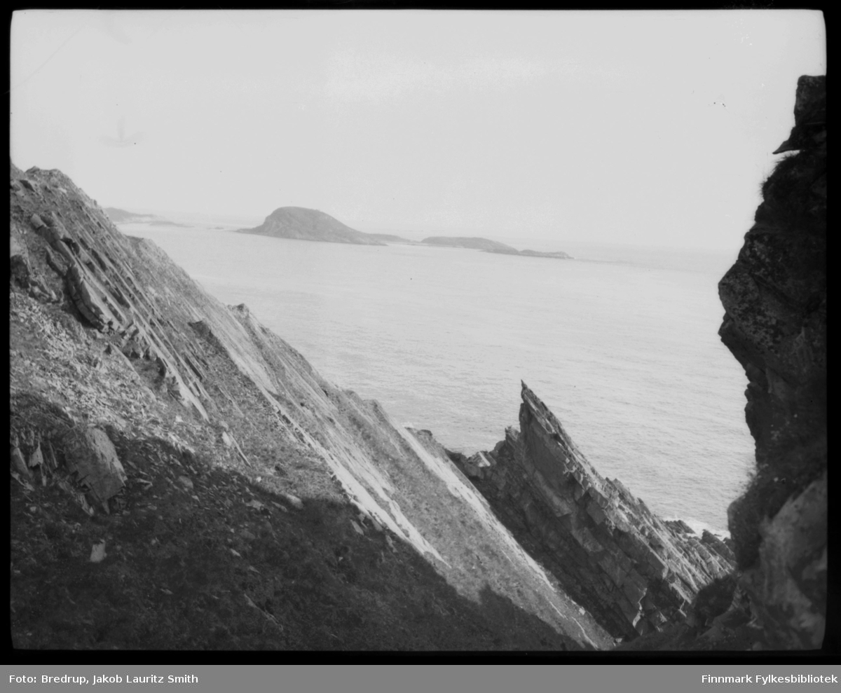 Fotografen har stått mellom noen klipper og tatt et oversiktsbilde utover noe som kan være Kongsfjord med Veidnesodden og øyene Kongsøya, Skarvholmen og Helløya.