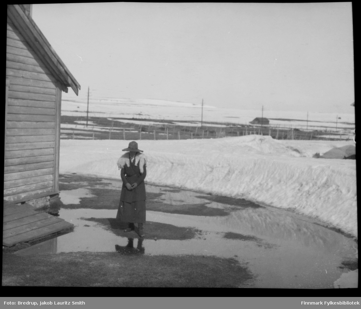 En dame kledd i hatt, drakt og med pelskrage står usikker og tripper foran store søledammer i vårløsninga.  Antakelig er bildet tatt i Vadsø.  I bakgrunnen et uthus, til venstre i bildet en annen bygning.