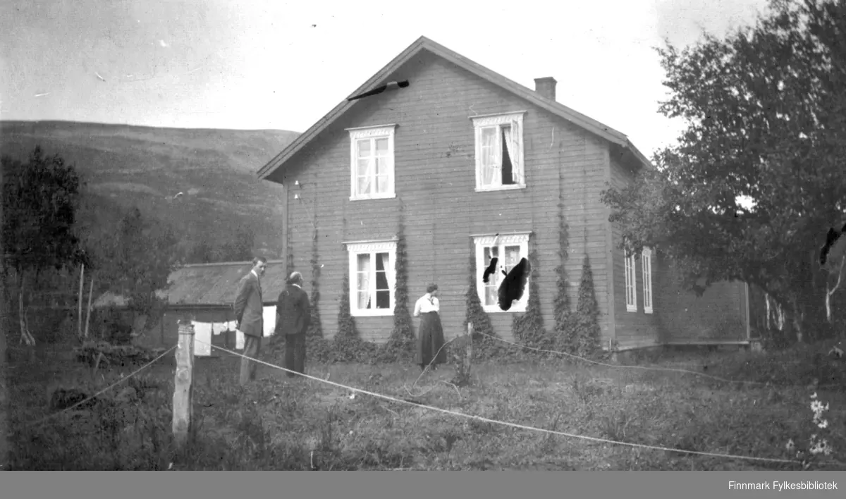 To menn og en dame står utenfor et hus i Rassejåkka i Tana en fin augustdag i 1916. Humle pynter veggen. Det var nok en nytteplante. I bakgrunnen henger det klær til tørk på en snor. På venstre siden av huset ligger det et uthus.
