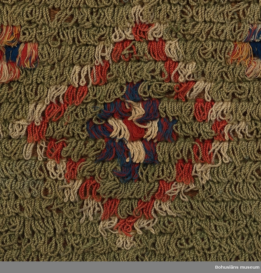 Täcke, prydnadsrya, troligen bröllopstäcke, vävd med inslagna nockor över hela ytan bildande ett grafiskt komponerat mönster i grönt, rött, blått och vitt med mittspegel och kantbård. Centralmotiv på blå botten föreställande två vita kannor med blå (mannen?) respektive röd (kvinnan?) botten med pip och handtag på en rektangulär uddkantad röd platta, på ömse längsgående sidor omgiven av en högrest blomsterstängel. Ovanför plattan en rad med omväxlande enkla och dubbla trianglar (timglas) i rött, grönt och vitt krönta av svårläst signering och datering, troligen "iis hmd 1858" i röda nockor. Nedanför centralmotivet en rad med upprepade kvadrater och trianglar (timglas) i omväxlande färger och på raden nedanför dessa en rad med rosor av rött och vitt garn på blå botten. 
Kring centralmotivet löper en 25 cm bred kantbård med romber på grön botten varannan romb har haft svarta nockor som nu helt är borta. (Se nedan). 
Överdelens hörn/yttersta kvadrater är prydda av en stjärna på röd botten. Nederdelens hörnen/yttersta kvadrater är prydda av ett rutmönster av röda och vita kvadrater samt  idag "nakna" kvadrater utan nockor där tidigare svarta nockor suttit. (Se nedan).

Bottenväven består av ofärgat, ljusbrunt entrådigt ullgarn i inslagsrips med varp av lintråd, den översta fjärdedelen av ett mörkare brunt ullgarn. Ryan består huvudsakligen av ullgarnsnockor av en enfärgad, tvåtrådig, ouppklippt ögla och två avklippta  ändar.  I vävnaden finns också andra varianter där bomullsgarn ingår; ett antal nockor består av tvåtrådigt ullgarn och entrådigt bomullsgarn i flera färger, blandade,  ett antal nockor består av entrådigt enfärgat bomullsgarn och dessa  ofta är centralt placerat i motivet samt ett antal  flerfärgade nockor av enbart bomullsgarn. Centralmotivets blå platta omges t. ex. av en bård helt och hållet i bomullsgarn.

De röda och gröna färgerna är växtfärgade, den blå troligen färgad med preussiskt blått med några smärre inslag av indigofärgat blått garn. Den svarta färgen har troligen erhållits genom efterbehandling med olika metallföreningar på ett mörkt växtfärgat garn. Denna behandling har gjort ullfibern spröd. Efter hand har nockorna frätts bort.

Ryan är  vävd i två våder, hopsydd med kaststygn på mitten. Den övre kanten är enkelt invikt och fållad och på baksidan förstärkt med fastsydd metalltråd med öglor för upphängning på vägg. Metalltråden är deformerad, vågig, genom tyngden av vävnaden. Underkanten är enkelt invikt och fållad. Långsidornas kanter är bitvis uppfransade.

Förvärvstillstånd: Relativt sliten och  blekt, bitvis trasig, smutsig.