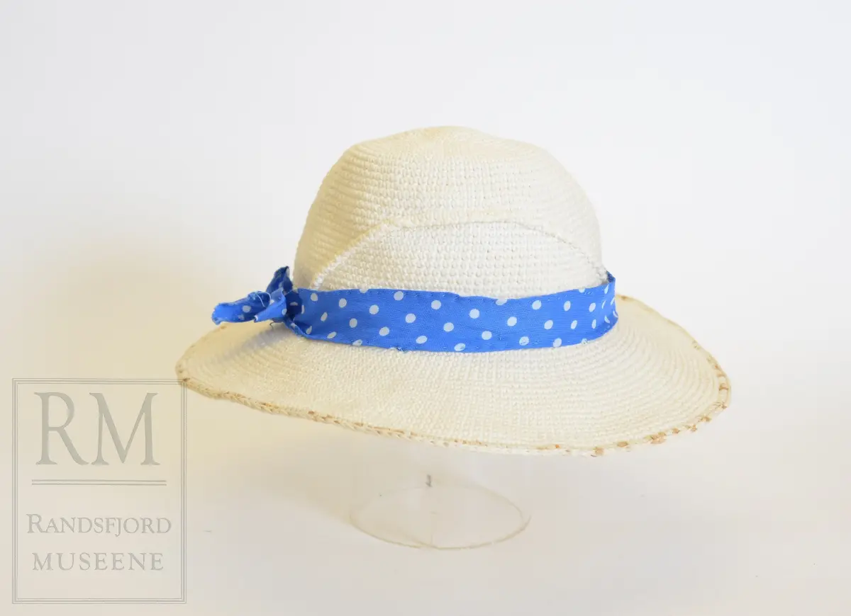 Sommerkjole m/hatt. a) Bomullstoff, blått med hvite prikker. Sammensydd i livet. Overdelen har Lukning foran med 5 hvite knapper og knapphull, puffermer. Krage med heppekant. 6 Bredder i utskrådd skjørt. Lukning til venstre for midt foran. 
b) Belte med knapp og knapphull. 
c) Hatt, Heklet av hvitt bomullsgarn. Ståltråd ytterst i Bremmen (rusten) Figurkant på pullen. Pyntebånd av kjoletøyet.