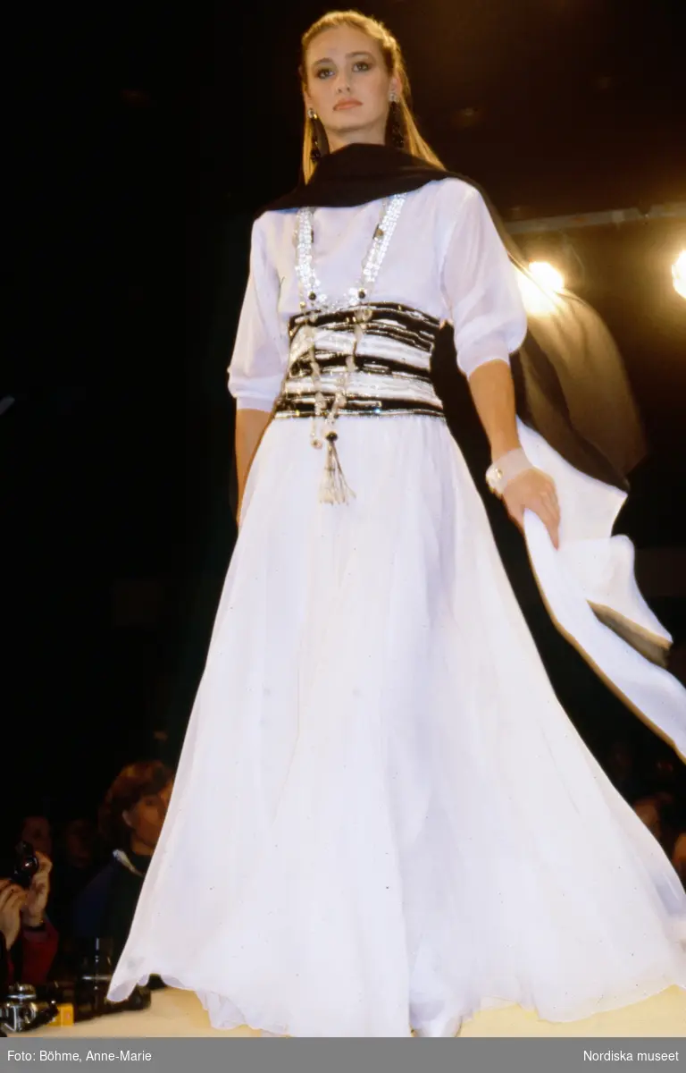 Modevisning. Modell i vit långklänning med randigt midjeband, svart sjal i halsen och halsband. Från Dior.