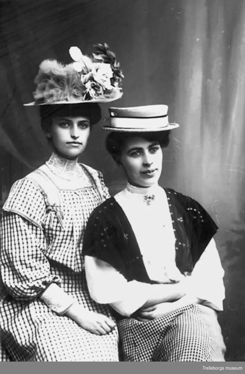 Två kvinnor, en i rutig klänning med hög spetskrage och hatt prydd med plymer och blommor och en i rutig kjol, vit blus och smalbrättad hatt garnerad med band.