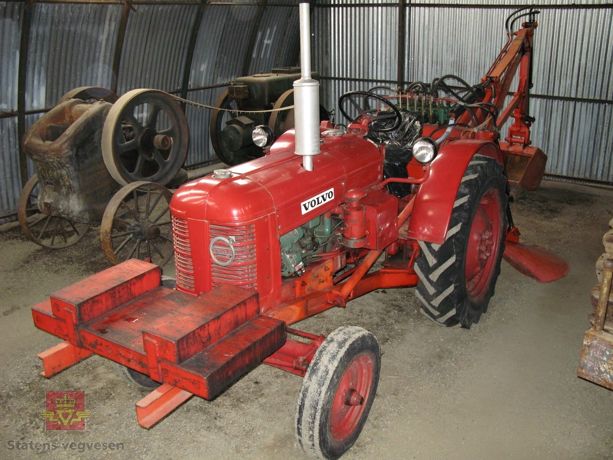 Volvo T 25 traktor med Hymas type 1 graver. Rød, svart og grønn traktorgraver lagd i hovedsak av metall, gummi og glass. Merket av produsentene. Dette er en jordbrukstraktor som er påmontert en graver, (gravemaskin), i bakkkant. Graveren er hydraulisk  drevet og har skuffe og to støtteben. Traktoren er 2-akslet har 4 hjul. Motoren er av typen C 22 B, som betyr en firesylindret firetakts bensinmotor med slagvolum 2,2 liter. Motoren yter 31 Hk. ved 2000 omdr./min. Motoren har tenningsmagnet med impulskobling. Girkasssen har fem gir fremover og ett gir bakover. Traktoren har 6 V elektrisk anlegg.  Ved ankomst NVM viser det seg at traktorens framdekk er meget sprukket, og støttebena til gravemaskina er festet med jekkestropper for å holde seg oppe. Maskina har også dårlige hydraulikkslanger.
