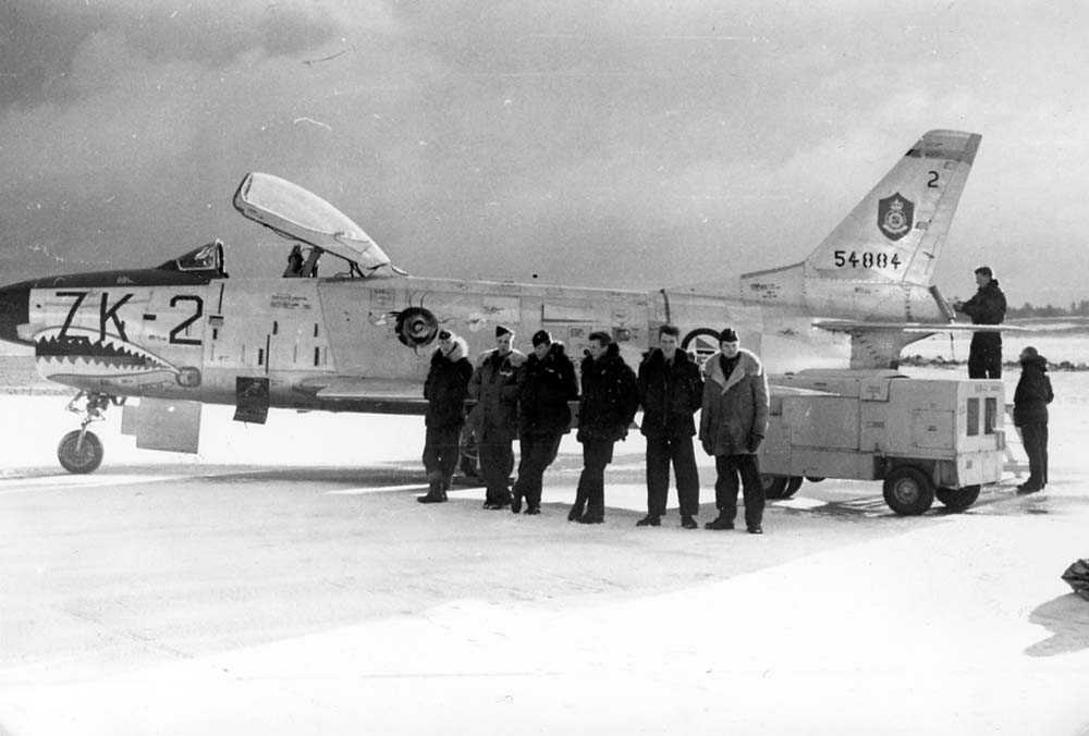Lufthavn. Ett fly på bakken. Seks personer står foran flyet, to personer står bak ved motoren på flyet. F-86K
