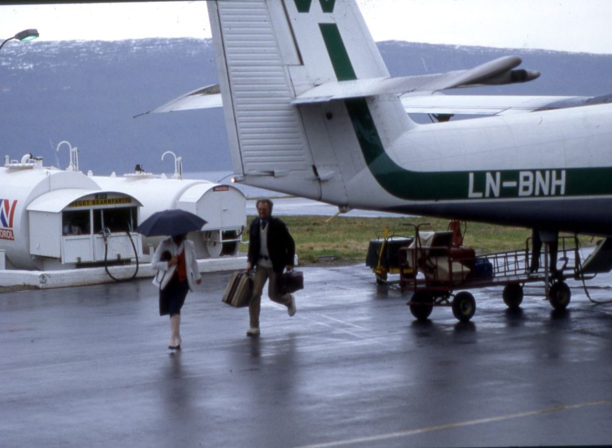 Lufthavn (flyplass). Et fly, LN-BNH, DHC-6-300 Twin Otter fra Widerøe parkert.. To personer (passasjerer) på vei fra flyet. Bagasje hentes ut av flyet. I bakgrunn tankanlagget.