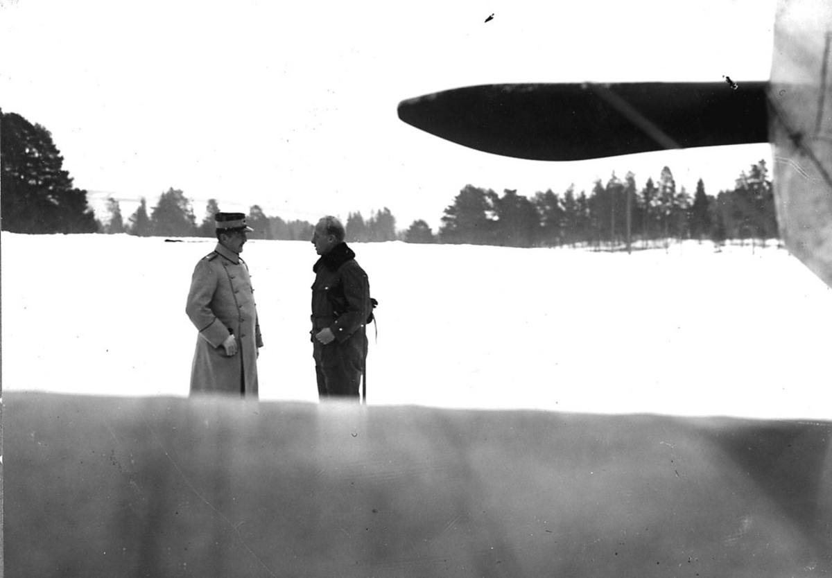   To personer. Svensk flyoffiser i samtale med norsk løytnant. Snø på bakken. Litt av ett fly sees i høyre billedkant.
