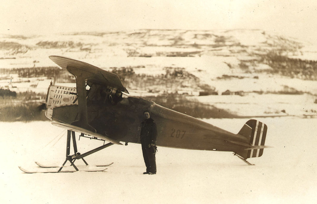 Åpen plass,  en person foran ett fly på bakken,  F.F.7 Hauk nr 207  med skiunderstell. Snø på bakken.