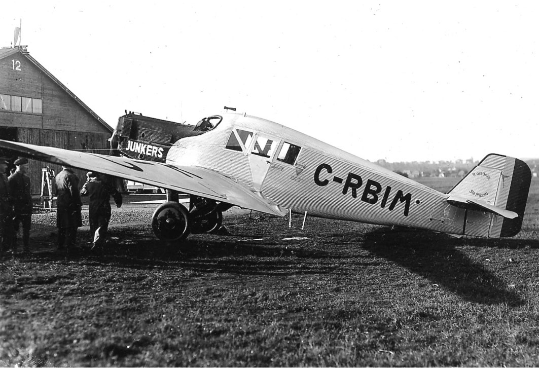 Åpen plass, lufthavn, ett fly på bakken, rumensk Junkers F.13, C-RBIM. trepersoner ved flyet. en bygning med 12 i røstet bak.