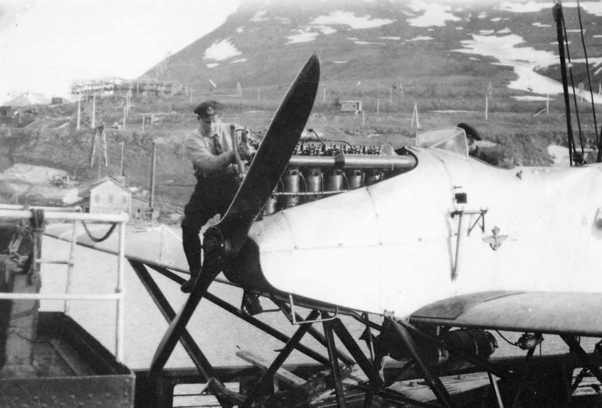    To personer, ettersyn på ett fly, som ligger ved land i havneområde. Svalbard.  Forparten på flyet, med motoren i nærbilde. 