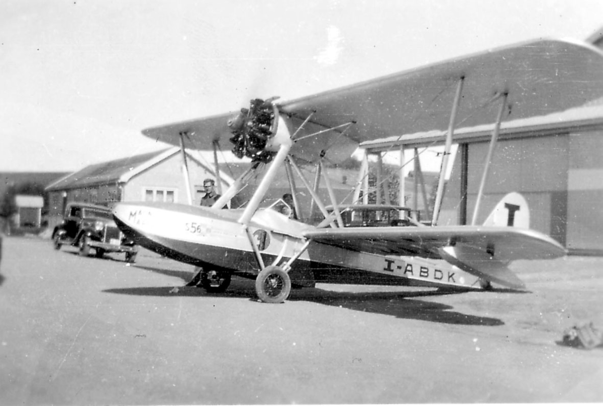 Åpen plass, ant. lufthavn, ett fly på bakken, Savoia   Marchetti S-56, I-ABDK. en person, en kjøretøy og to bygninger i bakgrunnen.