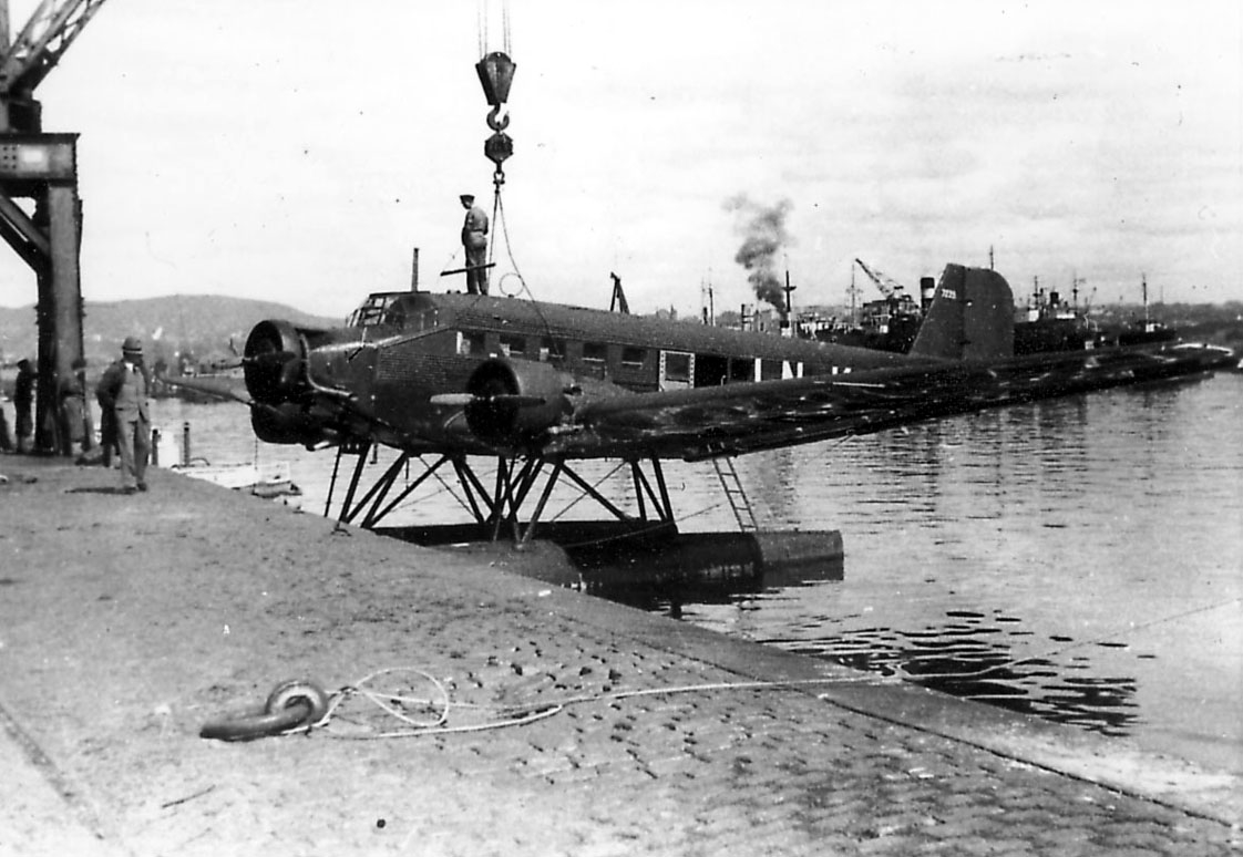 Ett fly, Junkers Ju 52/3mg7e LN-KAB  "Tyrihans", henger i heisekran. Kaidekke i forgrunnen. Røyk fra dampbåter bak.