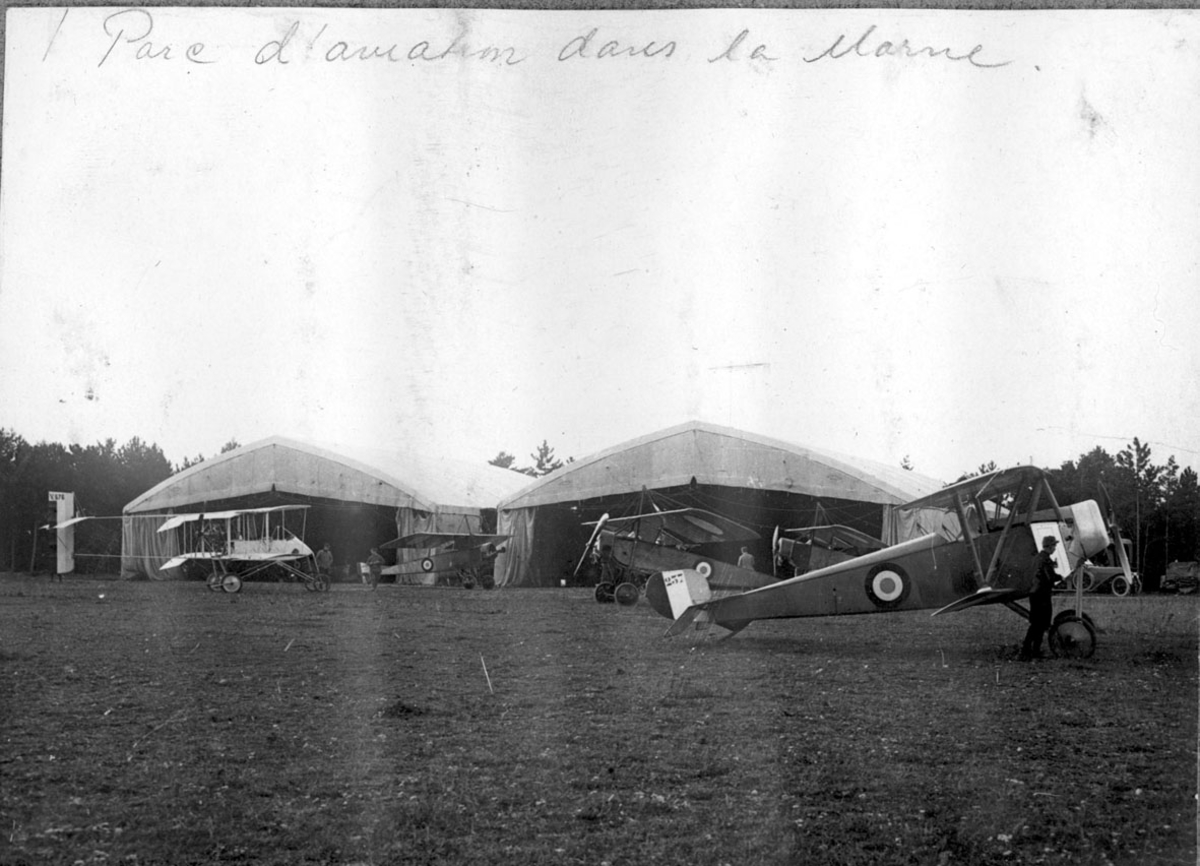 Flere fly på bakken. 1 stk. Voisin Biplane, 4 stk. Nieuport 11C.1. Flyene sett fra siden. 2 hangarer bak. 1 person foran.