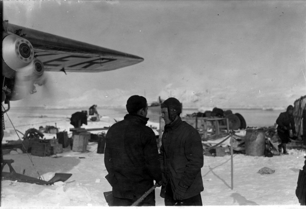2 personer i forgrunnen. T.v. flyvinge og en motor for propell, som er igang. Tilhører Fokker FVII "Josephine Ford". En del utstyr på bakken. Snø. Bak sees fartøy ute på havet, fjell.