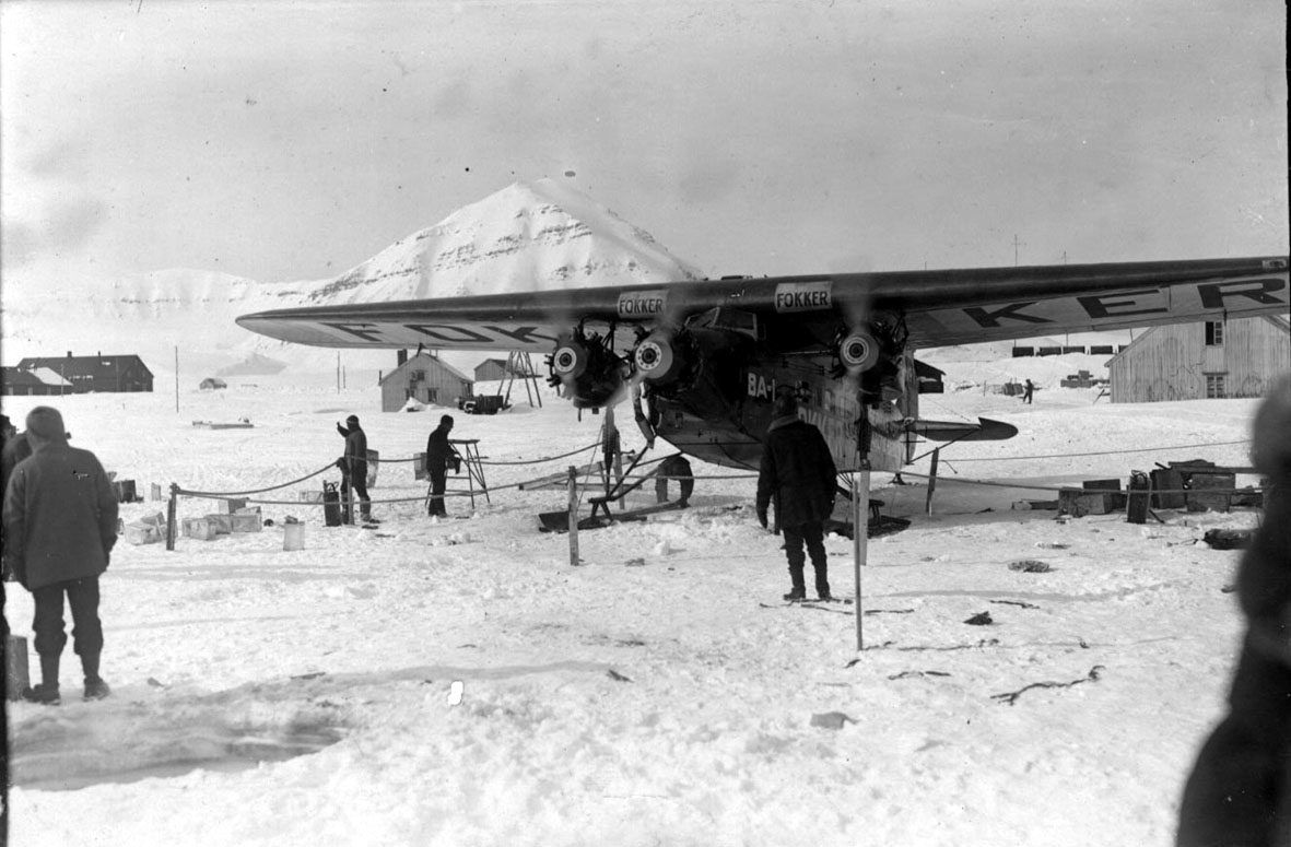 Fly, på bakken, Fokker FVII "Josephine Ford", med motorene i gang. Flere personer ved flyet. En del utstyr. Noen bygninger bak, fjelltopp.