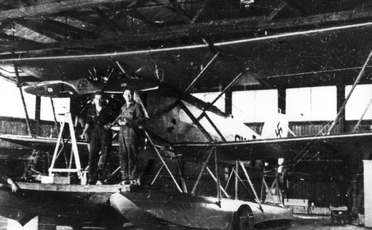 1 fly, MF-11, skrått forfra, inne i Marinens flyfabrikk. 3 personer oppstilt ved flymotoren. Hakekors på halepartiet. (Tyske kjennetegn).