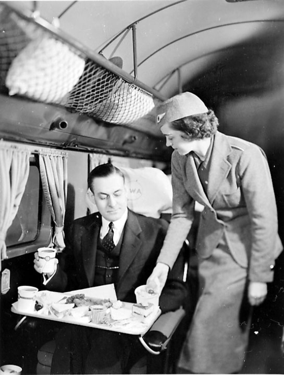 Inne i flycabinen på en DC-2. 2 personer. Flyvertinnen serverer mat til en passasjer.