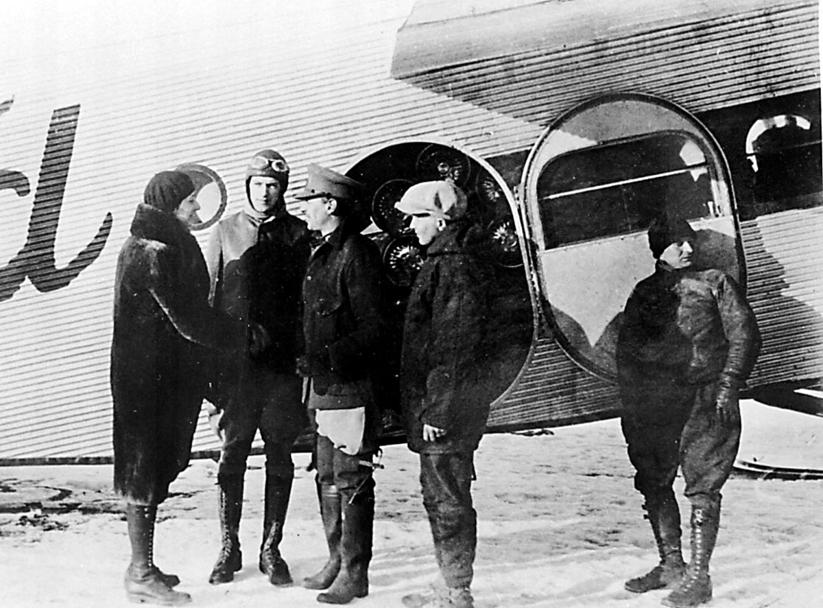 Portrett. 5 personer foran 1 fly, Ford. Snø på bakken.
1 kvinne hilser på "flymannskapet. Ant fra Byrds antarktiskekspedisjon med flyet "Ford"