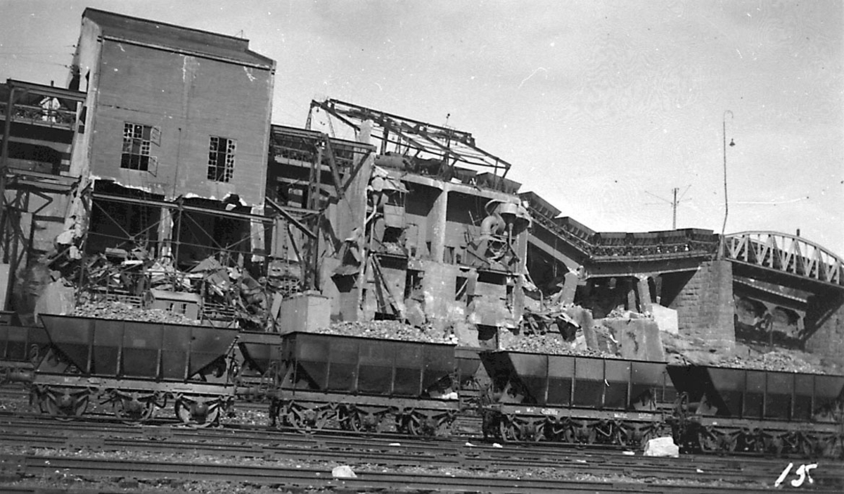 Tettsted/by. Jernbanevogner med malm i forgrunnen. Deler av jernbanekaia/industriområdet ødelagt av bombenedslag.