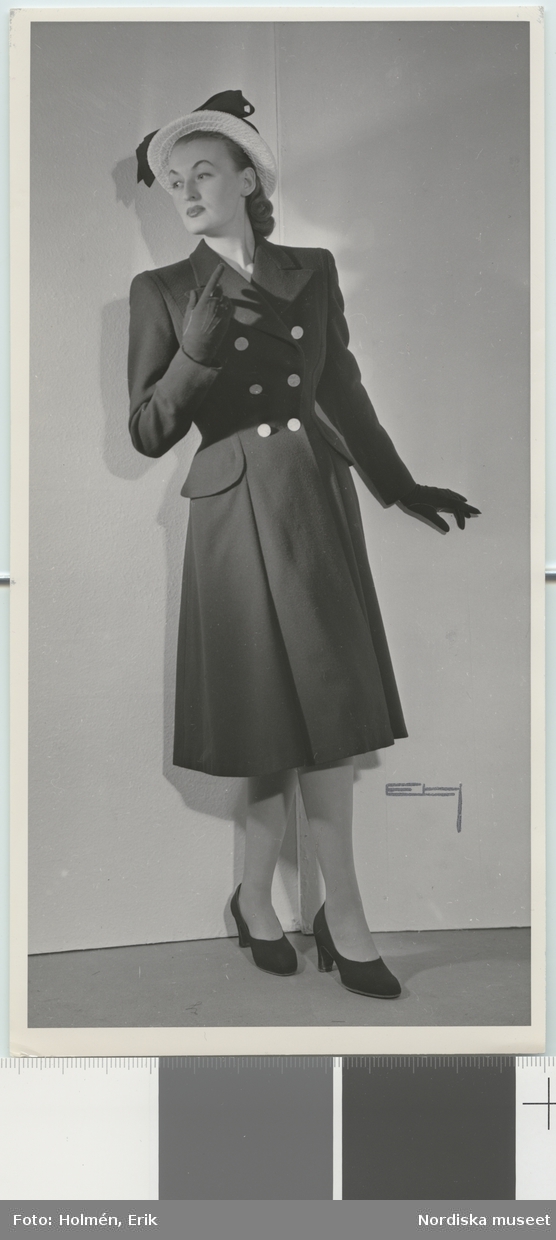 Nordiska Kompaniet. Kvinna i mörk kappa dubbelknäppt med vita knappar. Vit hatt med svart rosett. text på baksidan: "NK uppvisning mars 1947."