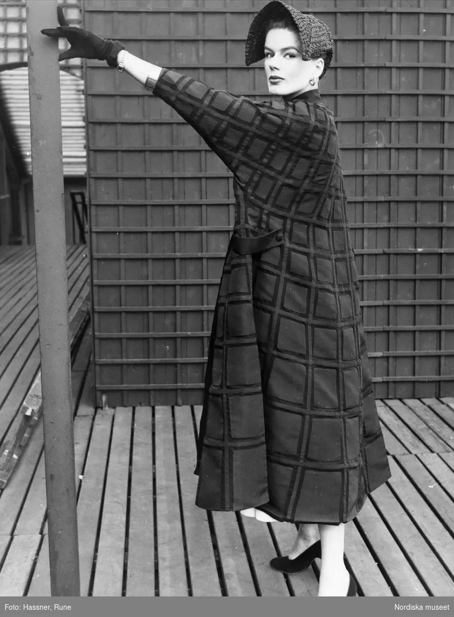 Modell klädd i rutig Fabiani- kappa, hatt, handskar och pumps. Står utomhus och lutar handen mot en stolpe. Italiana på Nordiska Kompaniet 1953.