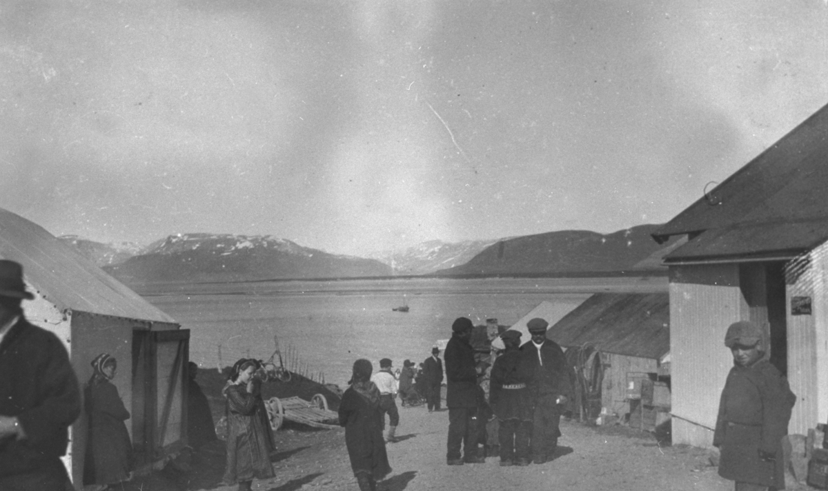 Markedet på Langnes 1925. Foran på bildet står en gutt, to jenter og tre menn på vegen. Bak på bildet flere mennesker, fjord og bakerst fjellene.