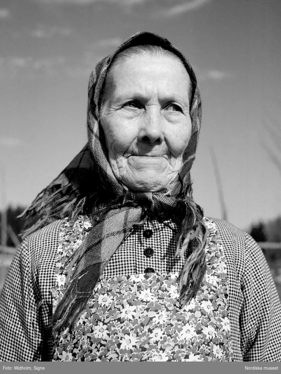 Porträtt av äldre kvinna med sjal på huvudet och blommigt förkläde.  "Brattsve Kristin" i Bergsjö socken, Hälsingland.