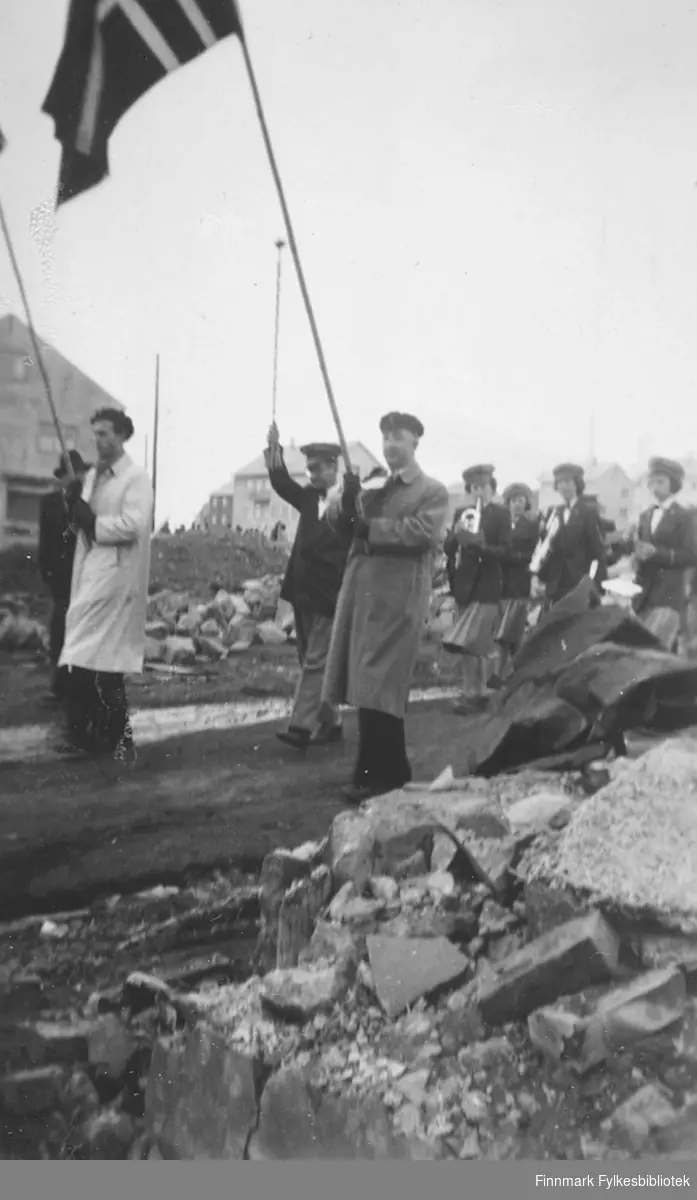 17. mai-toget i Vadsø sentrum, ca. 1950-1952. Per Bjørgan i lys frakk til venstre. I midten Olaf Holmen i uniform. Holmen var dirigent for kvinnekorpset som marsjerer bak. Mannen som holder fanen til høyre: Sigvald Foss, ansatt ved fylkesskattekontoret