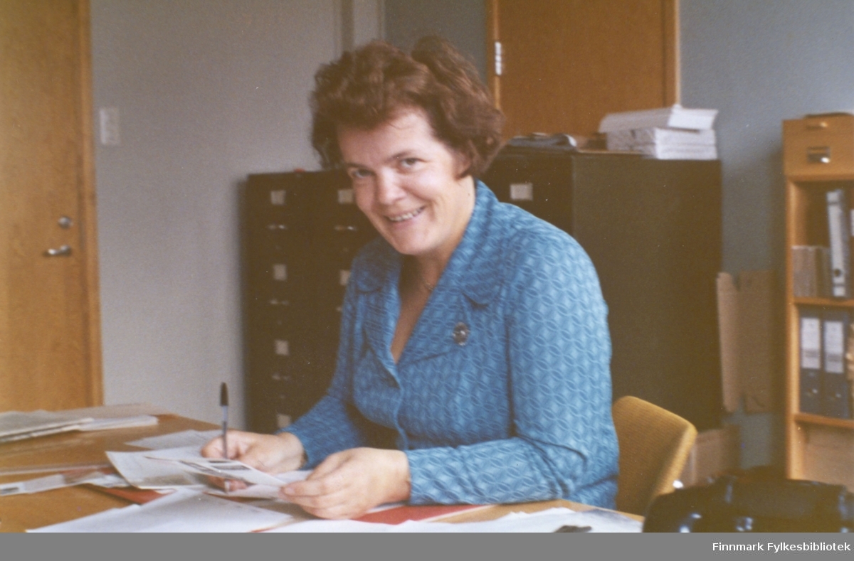 Ragnhild Ebeltoft på fylkesskattekontoret i Vadsø ca. 1973-1974. Ragnhild var ansatt ved fylkesskattekontoret i perioden 1951-1993. Bak henne ser vi arkivskap og permer i en bokhylle