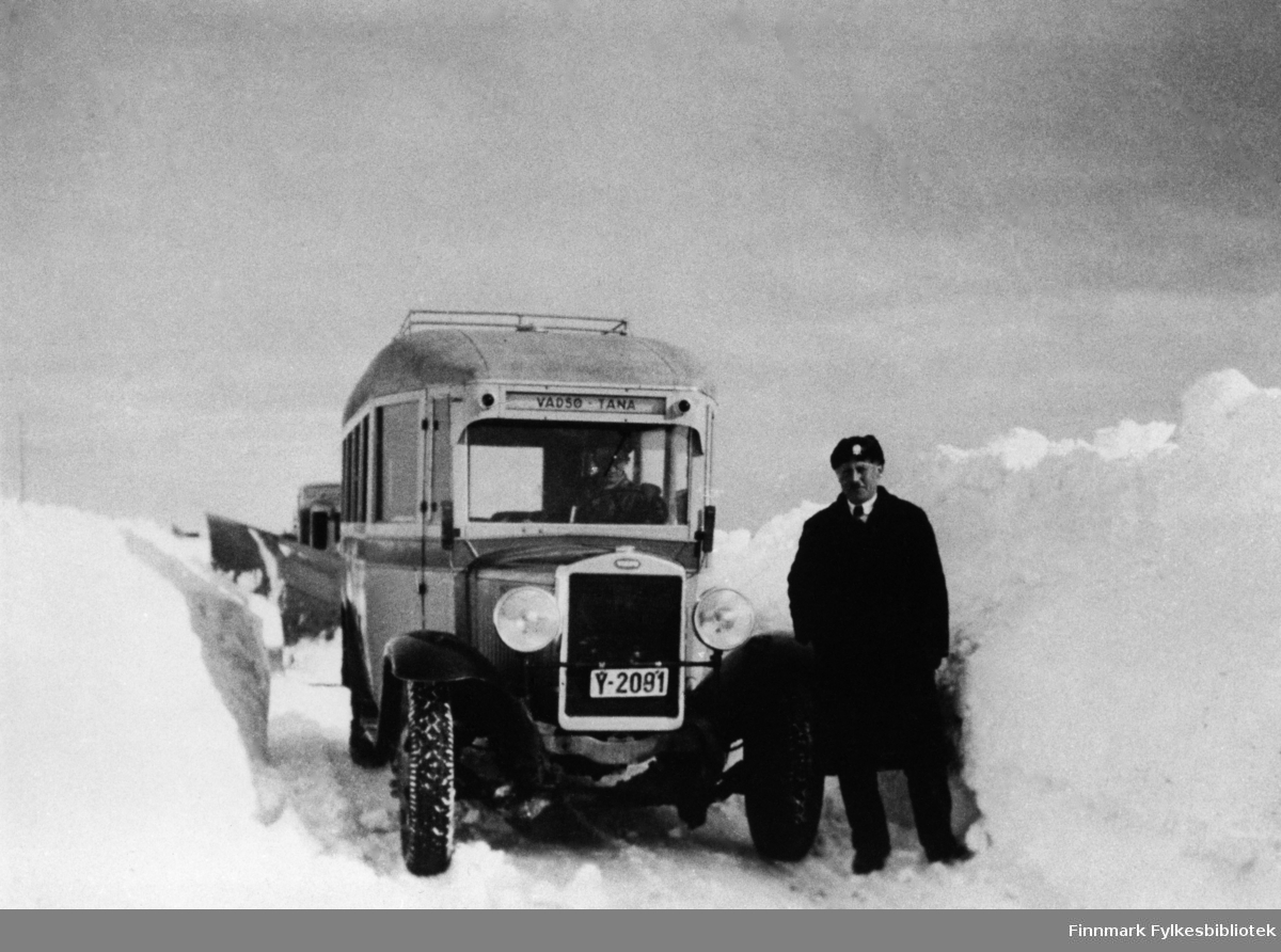 A/S Polarbil buss (y-2091, Volvo) brøyter veien, riksvei 950, Vadsø-Varangerbotn i 1940. Ved siden av bussen står en mann som heter Gustav A. Junge og en mann sitter i bussen (vi vet ikke navnet). I bakgrunnen ser man litt av brøytebilen. Se også bildene 577-588. A/S Polarbil var et rutebilselskap som eksisterte i tidsrommet 1920-1976. Ruteområdet dekte hovedstrekningene i Øst-Finnmark. Polarbil hadde hovedkontor i Vadsø. 1. januar ble selskapet med rutekonsesjoner, ansatte og bilmateriell overtatt av Finnmark Fylkesrederi og Ruteselskap (FFR).