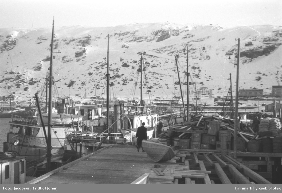 Vinter i Hammerfest havn. Store mengder bygningsmateriell, kasser og sandsekker ligger i stabler til høyre på kaia. Mannen midt på bildet er Ragnvald Jacobsen, iført mørke klær. Rett bak han ligger en liten trebåt. Flere båter ligger fortøyd ved kaia og noen brakker ses i bakgrunnen ved foten av fjellet Salen og i strandkanten. Fjellet er stort sett snødekt og sikk-sakkveien ses midt på bildet.