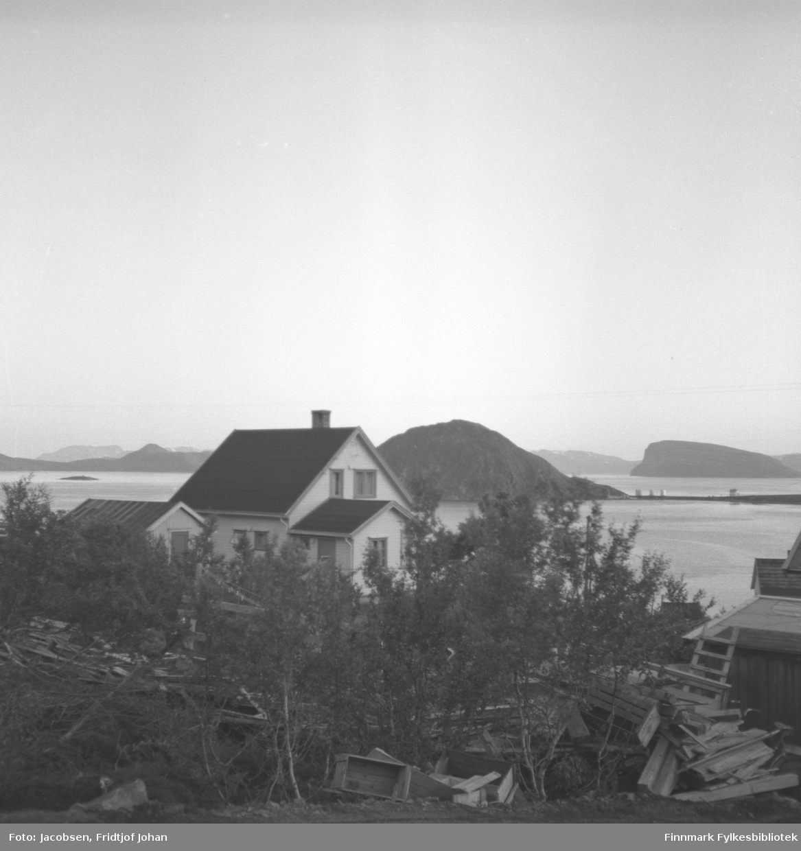 Utsikt mot huset til lærerinne Anna Andersen i Rypefjord, fotografert fra Arne Nakkes eiendom. Det står mange løvtrær foran på bildet og mye treskrot ligger på bakken som planker og kasser. På andre siden av Rypefjorden til høyre på bildet ses Essos tankanlegg på Rypklubbeidet med Rypklubben til venstre. Over tankanlegget ses øya Håja i Sørøysundet. Til høyre på bildet ligger øya Seiland med Rypklubbskjæret foran.
