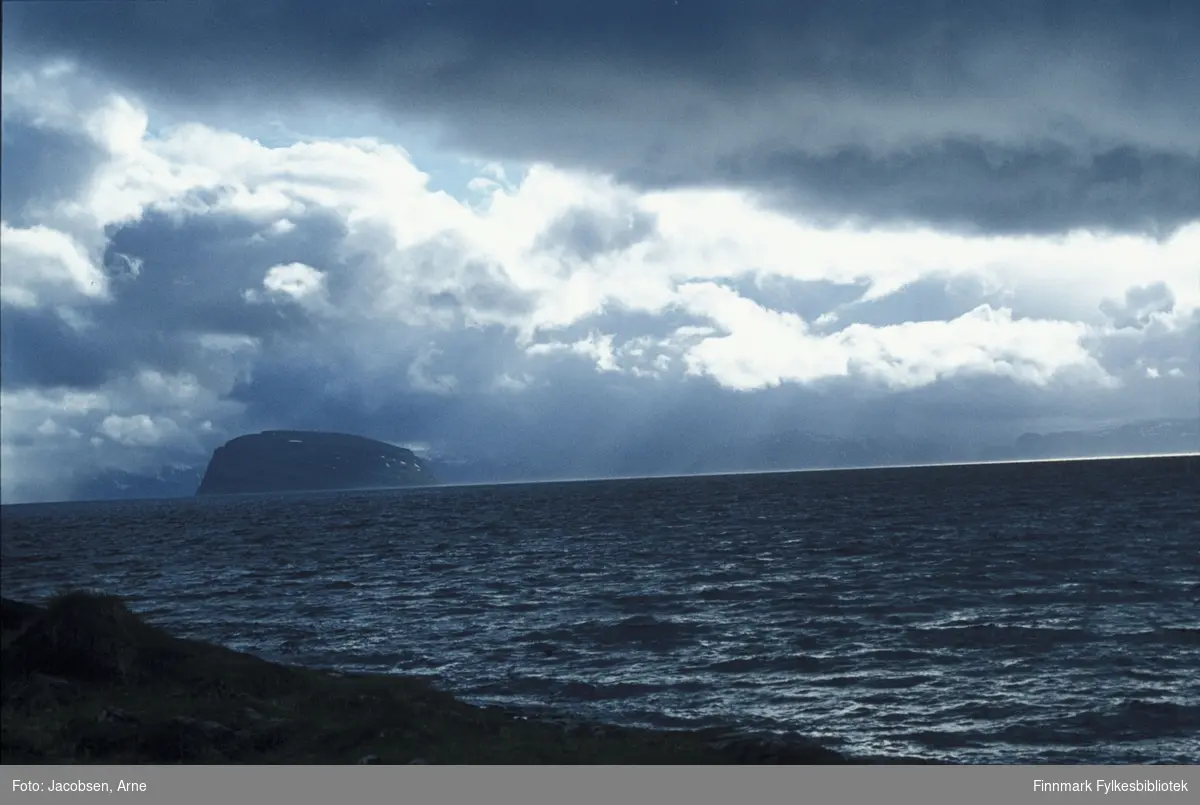 Den karakteristiske formen på øya Håja vises mot tunge skyer, sett fra Kvaløya. Sørøya skimtes såvidt i bakgrunnen. Noe vind, men oppholdsvær. Bildet er nok tatt en sen vårdag/tidlig sommer pga. snøflekkene som ligger igjen på øya.
