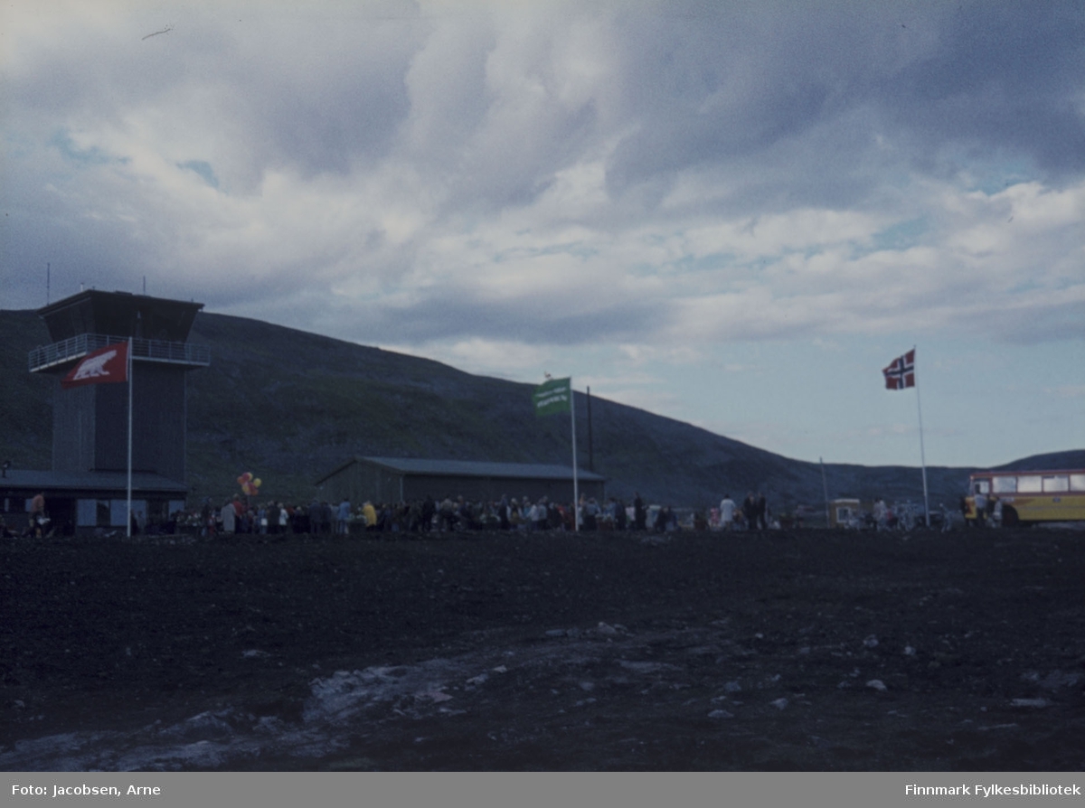 Hammerfest lufthavn åpnes 30. juli 1974. Tårnet til venstre og garasjeanlegg midt på bildet. Helt til høyre står en FFR-buss. Overskyet vær med litt vind. Mot tårnet ses Hammerfest-flagget med isbjørn-motivet og flagget i midten er Widerøes firmaflagg. Mye folk og ballonger, til høyre for tårnet, viser av det er feiring/markering.