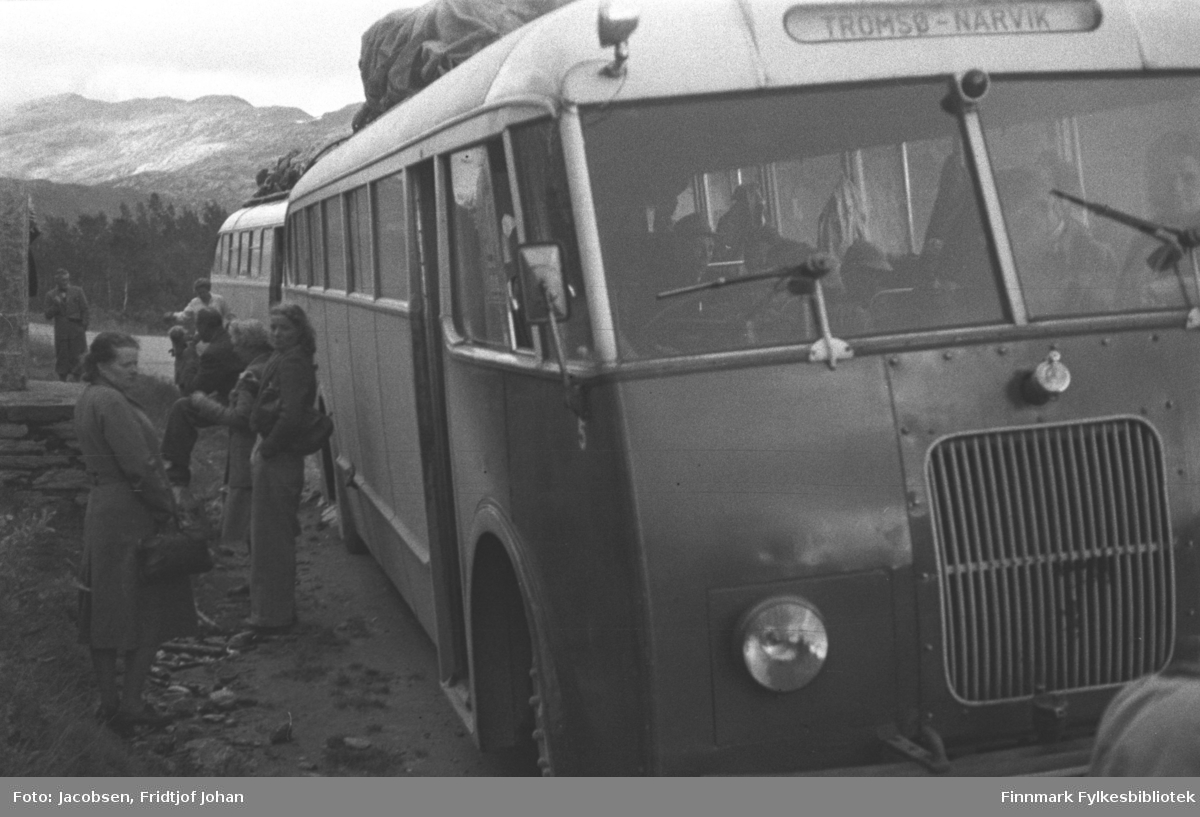 Bussen som gikk fra Tromsø til Narvik. Bussen tilhører antakelig TIRB - Troms Innland Rutebil. TIRB hadde langruter fra Narvik i sør og nordover til Tromsø og Alta. Strekningen Narvik-Alta var en del av Nord-Norge-Bussen, som hadde ruter mellom Kirkenes i nord og sørover til endestasjonen på Nordlandsbanen. I tidsrommet 1947-1955 var Lønsdal endestasjon og bussene kjørte med passasjerer og post videre nordover derfra. 1955-1958 var Røkland endestasjon, så ble det åpning til Fauske i 1958 og til Bodø i 1962. Her står den på grensa mellom Finnmark og Troms fylke. Noe av grensesteinen ses helt til venstre på bildet. Damen i kåpe til venstre i bildet er muligens Betty Knoblock. Noe skog ses i terrenget bak bussen og et høyt fjell ses oppe til venstre.