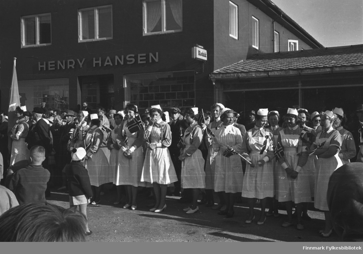 En gruppe sykepleiere i uniformer, samt noen andre mennesker står ved Henry Hansens fotoforretning og feirer med flagg i hendene. Dagen er solrik og det er sommer.