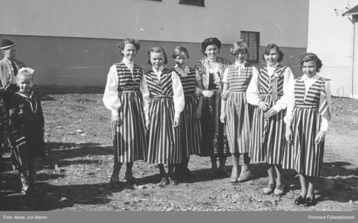 17. mai i Øksfjord, 1954. Flere av jentene ved skolen hadde sydd like drakter i håndarbeidstimene på skolen. Her står de sammen med læreren sin foran samfunnshuset i Øksfjord