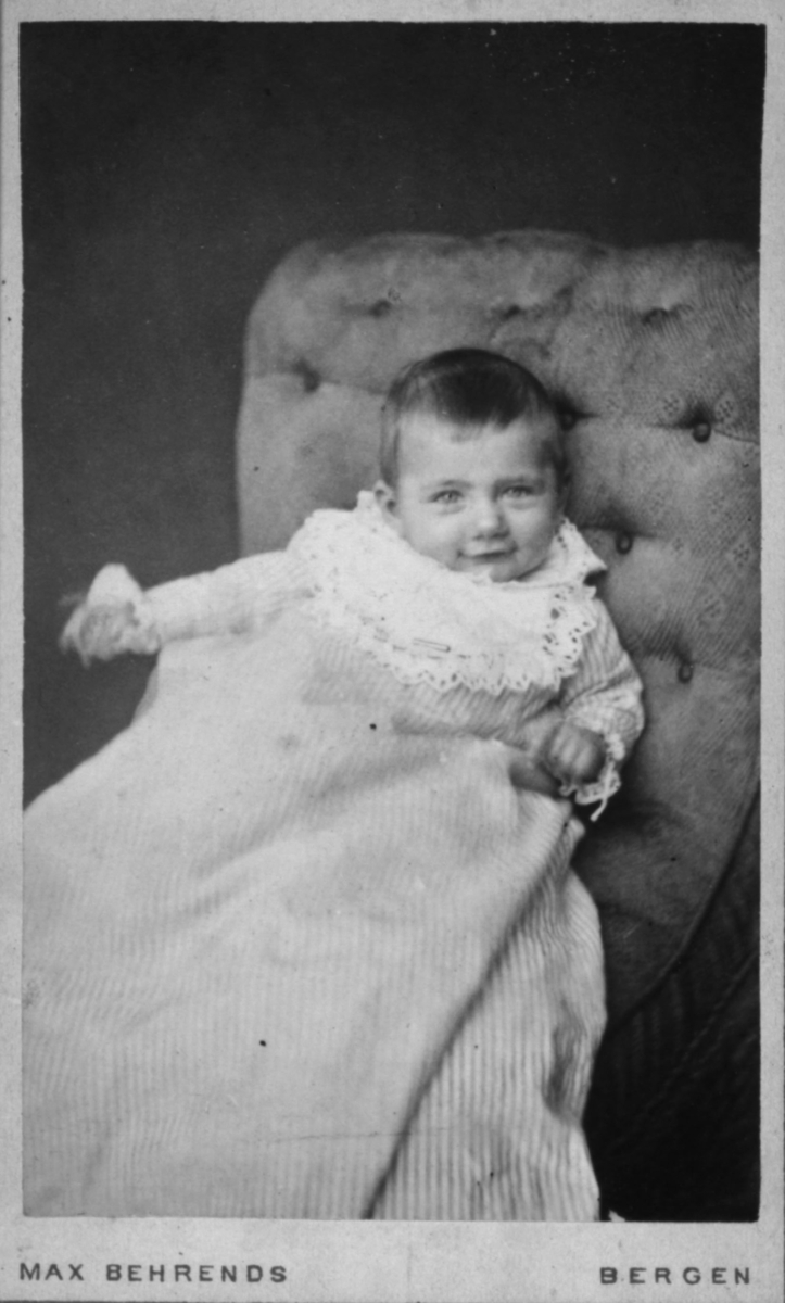 Portrett av et blidt barn i lang kjole - det er vanskelig å si om dette er ei jente eller en gutt. Barnet ser ut til å være 1-2 år