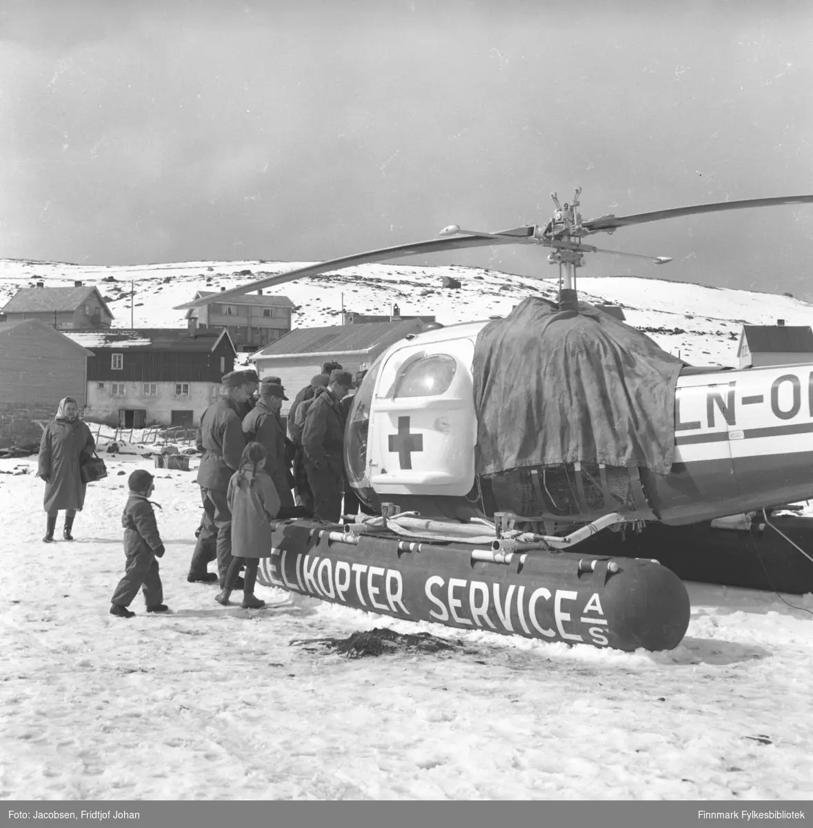 Et helikopter fra Helikopter Service A/S står på Hammerfest stadion med en del menn i militærklær, noen barn og en kvinne rundt. Bebyggelsen i Skytterveien ses i bakgrunen.