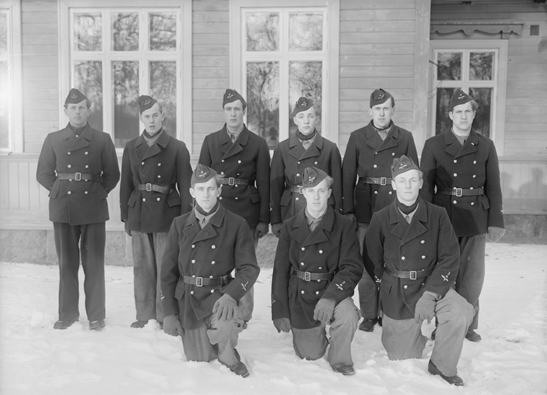 Elever vid vicekorpralskolan på F 3 Östgöta flygflottilj, 1940-1941. Nio stycken elever. Grupporträtt framför byggnad, vintertid.